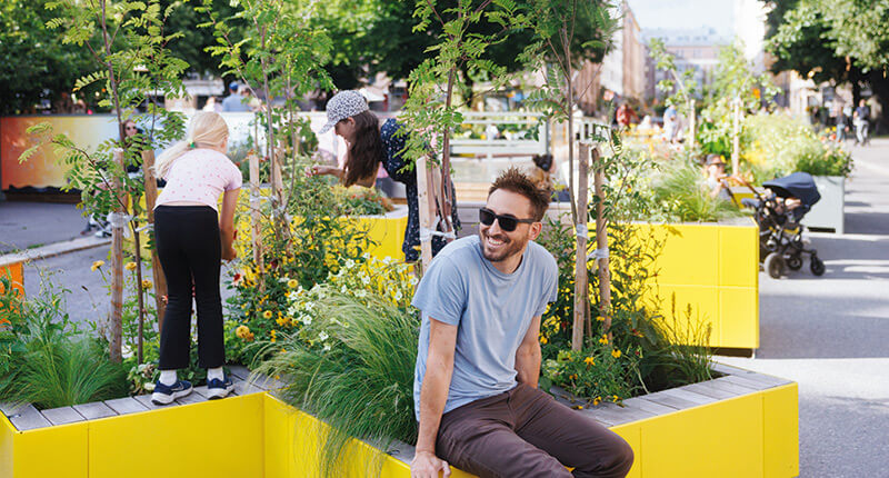 Mehrere Menschen nutzen eine gelbe Sitzinsel mit integrierten Pflanzen in einer urbanen Straße