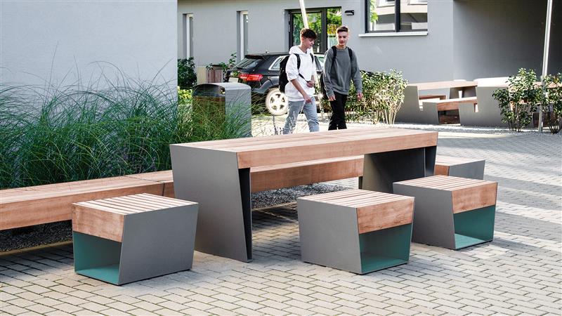 Sitzbank und Tisch bestehend aus Stahlteilen und Holzbelattung platziert auf einem Schulgelände, im Hintergrund sind zwei Jungs zu sehen