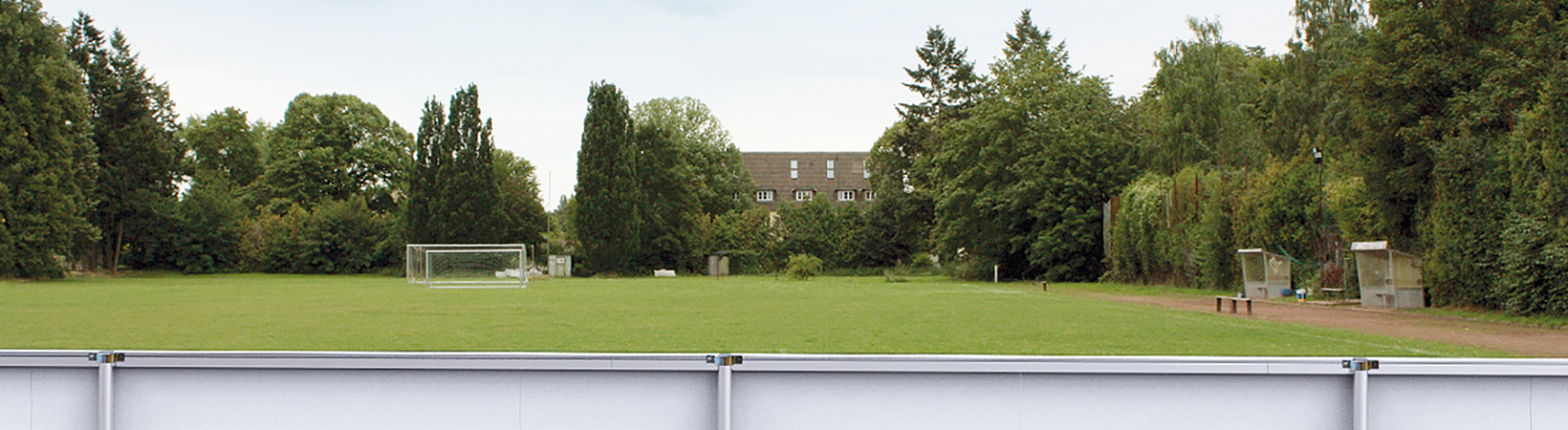 Leere weiße Werbebande vor einem Fußballtor auf einem Rasenplatz mit Bäumen und einem Gebäude im Hintergrund