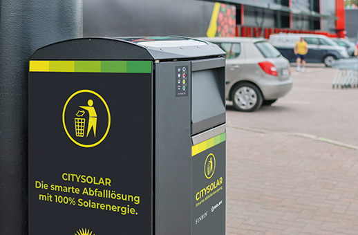 Abfallbehälter mit Aufschrift 'CITYSOLAR Die smarte Abfalllösung mit 100% Solarenergie' auf einem Parkplatz mit Autos im Hintergrund