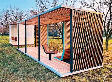 Offene Pergola mit Holzlatten an den Seiten und zwei modernen Sitzbänken auf Holzterrasse im Garten.