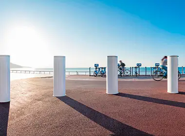 Mehrere weiße Hochsicherheitspoller stehen auf einem gepflasterten Platz am Wasser, im Hintergrund sind zwei Radfahrer zu sehen.