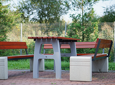 Freiluft-Sitzgruppe mit Tisch und zwei Bänken aus Beton und Holz auf gepflastertem Boden, umgeben von Grün und Zaun im Hintergrund.