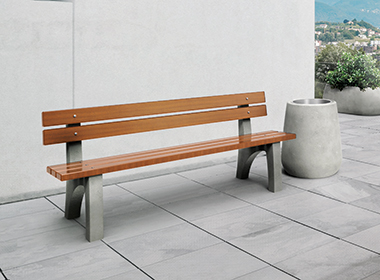 Sitzbank mit Holzlatten und grauen Betonfüßen auf gepflastertem Boden vor weißer Wand, daneben ein großer runder Pflanzkübel mit grünem Busch.