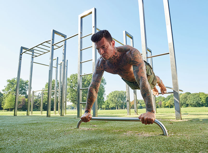 Mann macht Sport an einer Calisthenics Anlage auf einer Grünfläche mit Bäumen im Hintergrund