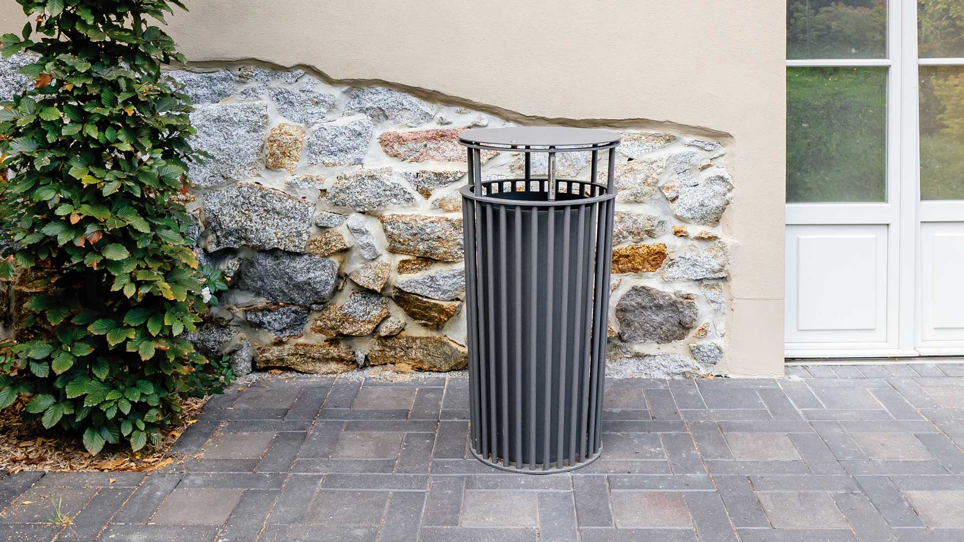 Runder Abfallbehälter aus Metall mit senkrechten Streben vor einer Steinmauer und neben einer Pflanze auf gepflastertem Boden