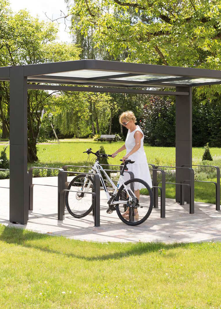 Frau in weißem Kleid sichert Fahrrad unter modernem Fahrradunterstand mit Flachdach in Parkanlage