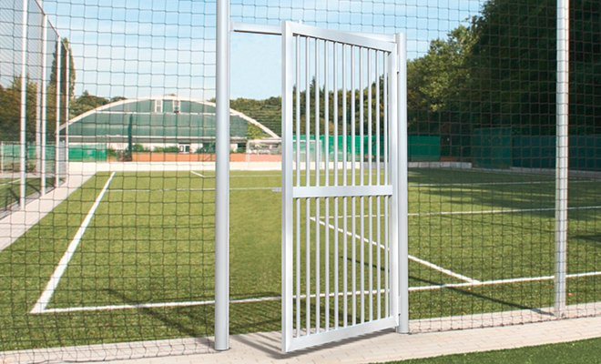 Offenes weißes Metalltor in einem Ballfangnetz an einem Fußballfeld mit Kunstrasen und Stadiontribüne im Hintergrund