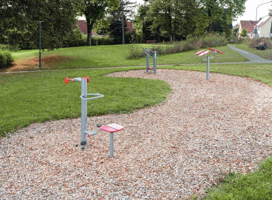 Mehrere Outdoor-Fitnessgeräte auf einer mit Holzschnitzeln bedeckten Fläche in einem Park