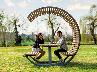 Zwei Personen sitzen sich an einem runden Holztisch mit integrierter, halbkreisförmiger Überdachung aus Holzlatten gegenüber, im Hintergrund eine Wiese und Bäume.
