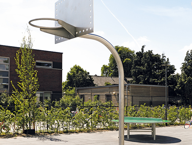 Freistehender Basketballkorb mit Metallpfosten und transparentem Backboard auf einem gepflasterten Platz vor Büschen und Gebäuden