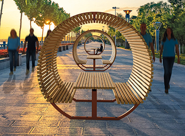 Mehrere runde, aus Holzlatten gebaute Sitzbänke in einer Reihe auf einem gepflasterten Weg mit Bäumen und Laternen im Hintergrund.