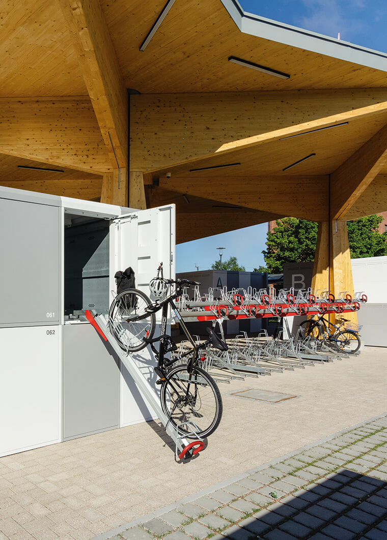 Fahrradgarage mit geöffnetem Schrank, in dem ein Fahrrad auf einer schrägen Rampe steht, und weiteren Fahrradständern unter einem modernen Holzvordach