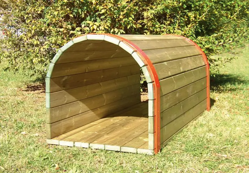 Halbrunder Hundetunnel aus Holz mit roten Metallstreben, auf einer Wiese vor Büschen.