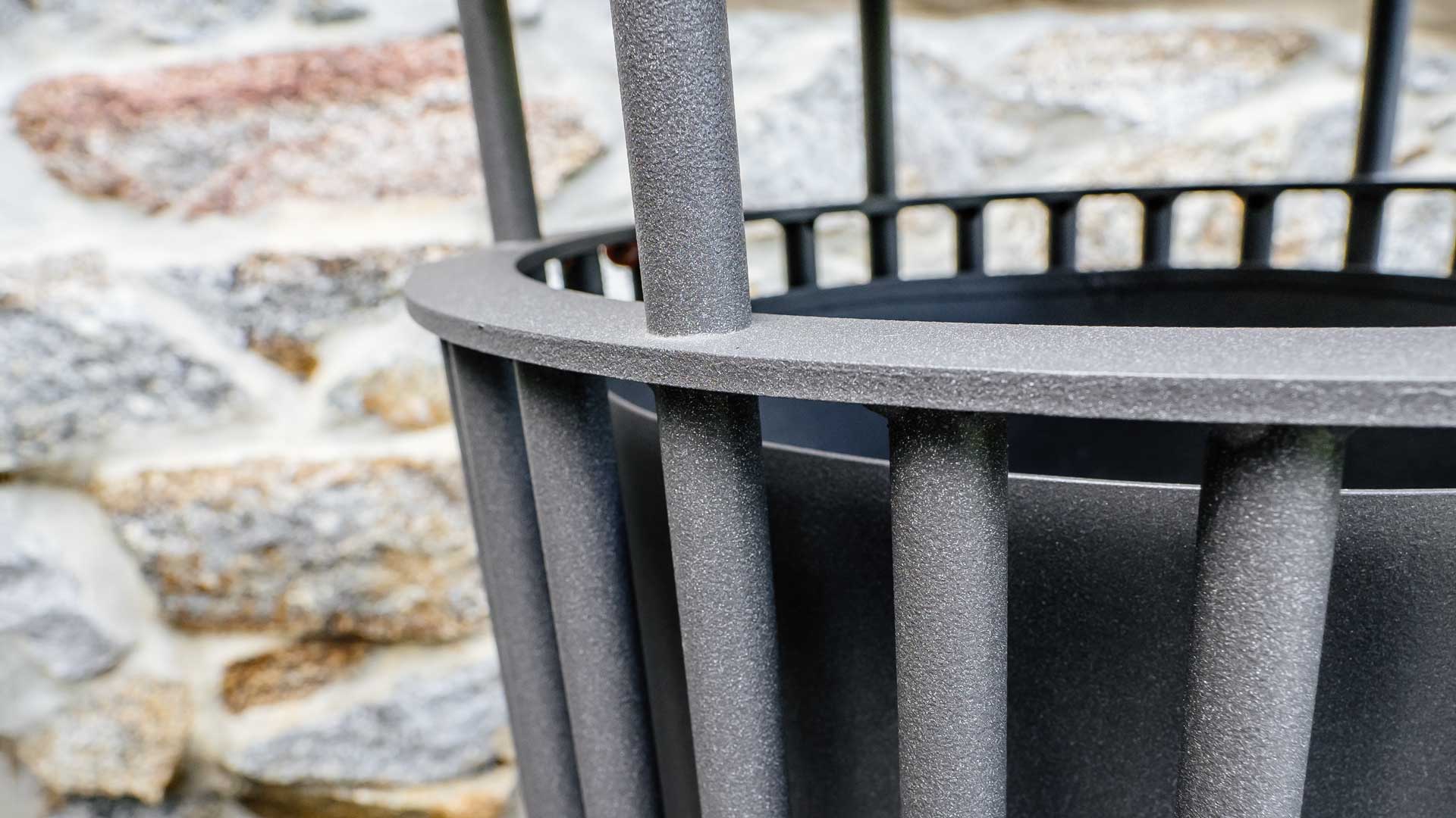 Metall-Abfallbehälter mit senkrechten Streben vor einer Steinmauer