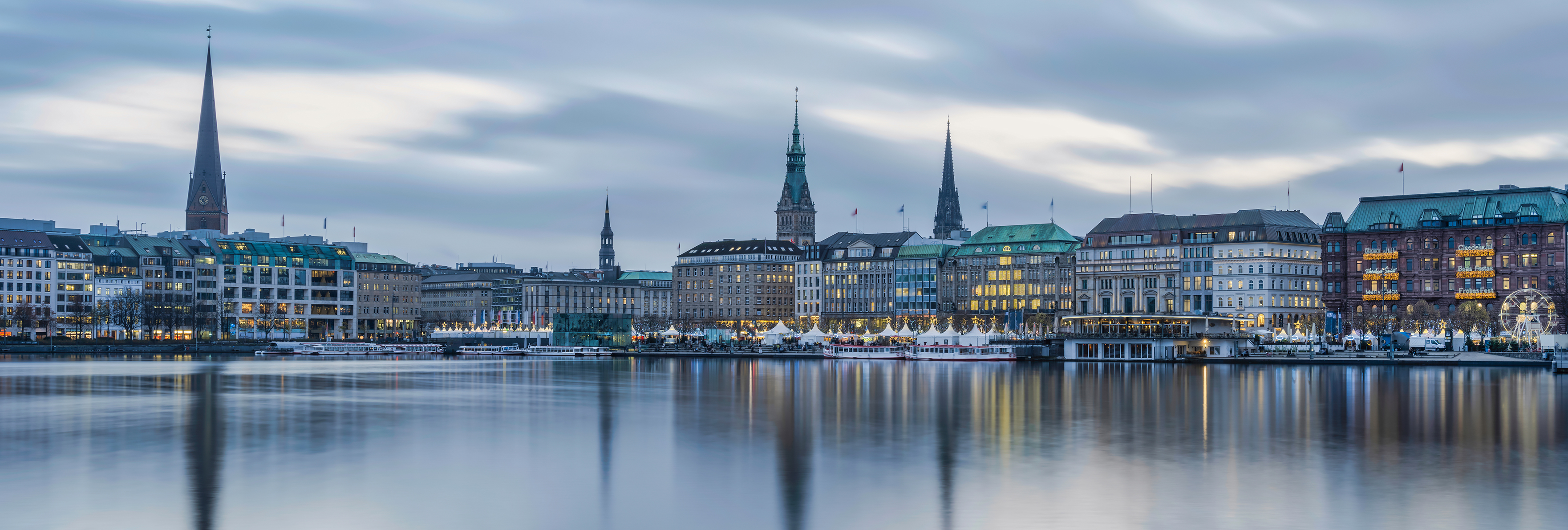 Panoramaaufnahme der Hamburger Innenstadt mit mehreren Kirchtürmen und Gebäuden am Wasser bei bewölktem Himmel.