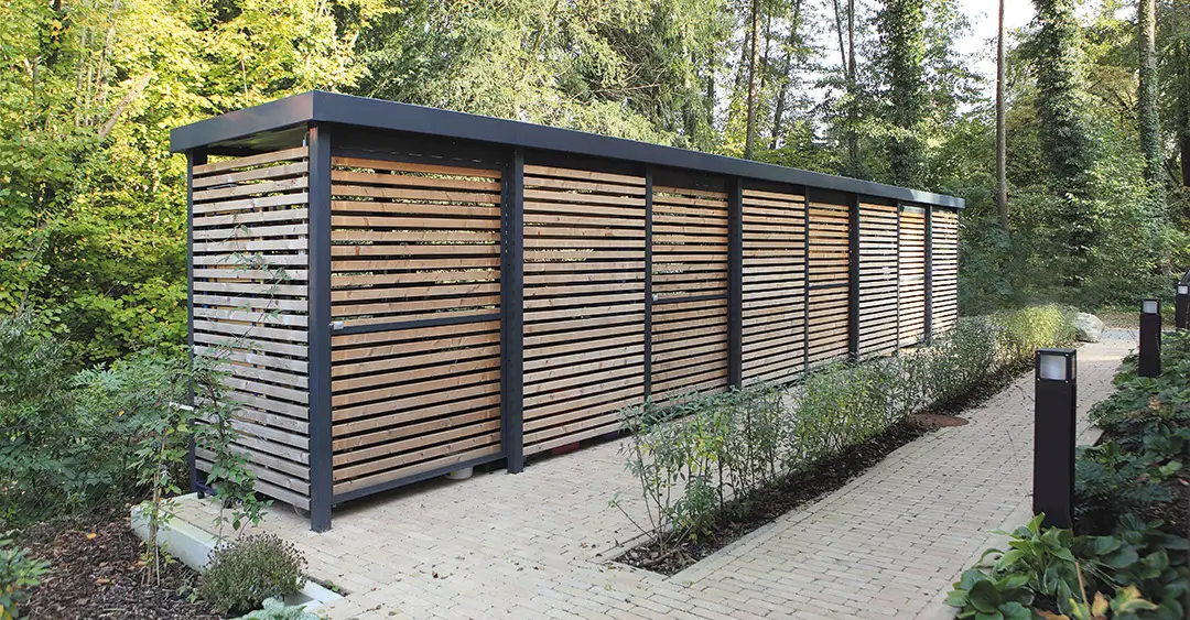 Längliche Überdachung aus Holzlatten mit schwarzem Metallrahmen neben gepflastertem Weg im Waldgebiet.