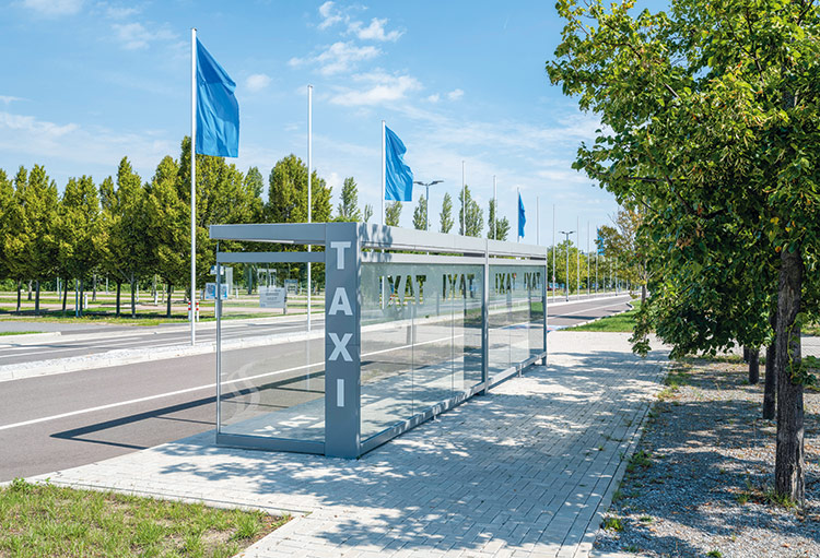 Glaswartehalle mit silbernem Rahmen und vertikalem Schriftzug 'TAXI' an einer breiten Straße mit blau wehenden Fahnen und Bäumen
