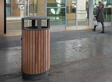 Runder Abfallbehälter mit vertikalen Holzlatten und schwarzem Metallrahmen auf einem Gehweg vor einem Gebäude.