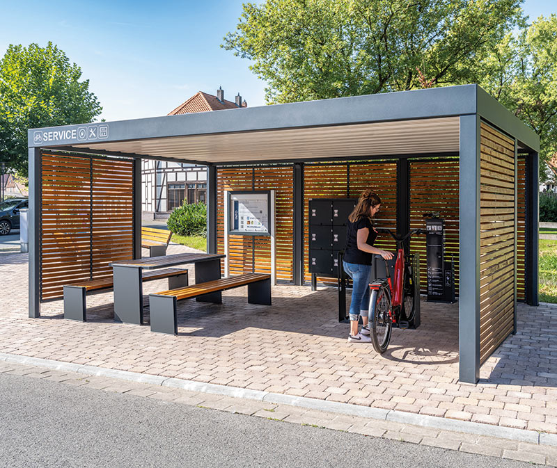 Überdachte Fahrradabstellanlage mit Sitzbank, Tisch und abschließbaren Fahrradboxen, umgeben von Holzlamellen und Metallrahmen