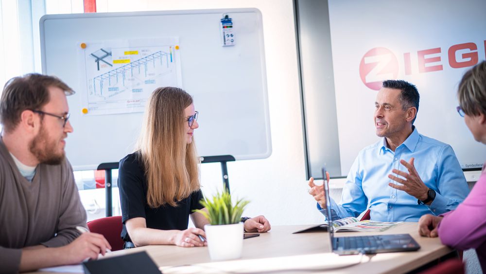 Vier Personen sitzen an einem Tisch in einem Besprechungsraum mit einem Whiteboard im Hintergrund und diskutieren