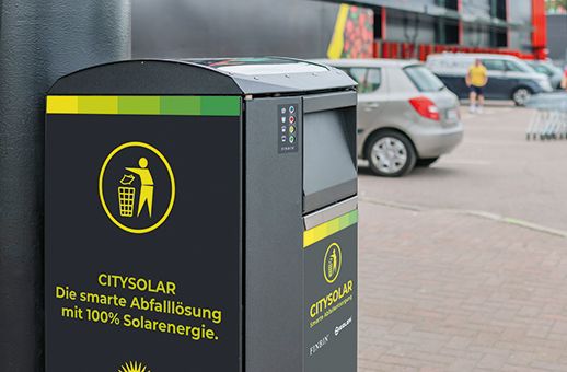 Abfallbehälter mit Aufschrift 'CITYSOLAR Die smarte Abfalllösung mit 100% Solarenergie' auf einem Parkplatz mit Autos im Hintergrund