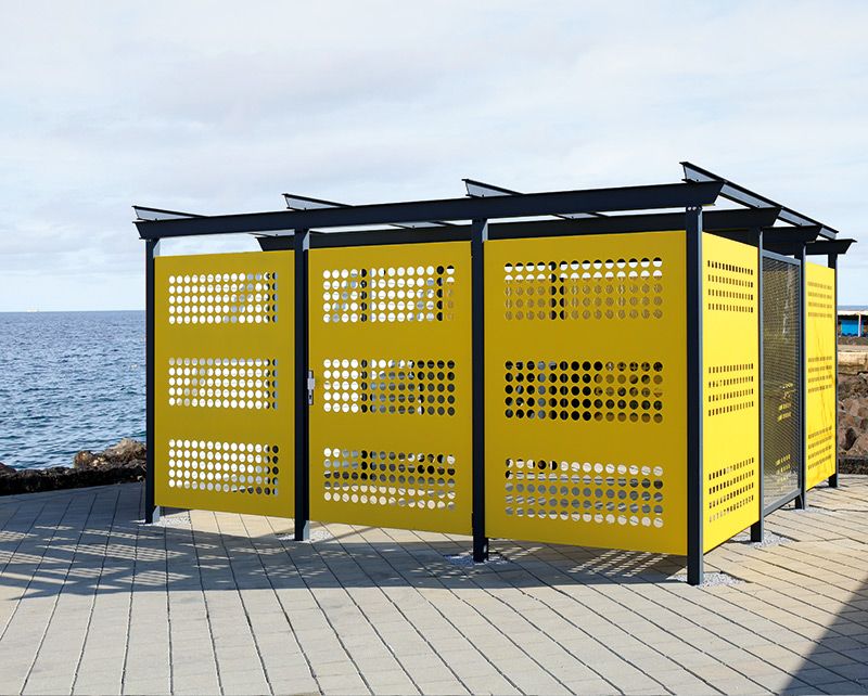 Gelbe Wertstoffumhausung mit Lochmuster und schwarzem Metallrahmen auf gepflastertem Boden am Meer