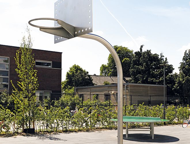 Freistehender Basketballkorb mit Metallpfosten und transparentem Backboard auf einem gepflasterten Platz vor Büschen und Gebäuden