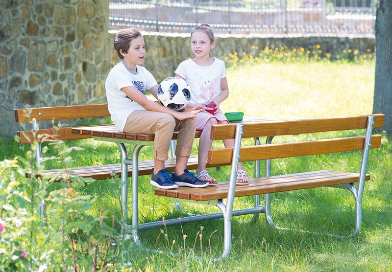Zwei Kinder sitzen auf einer Holzbank mit Metallgestell im Garten, eines hält einen Fußball