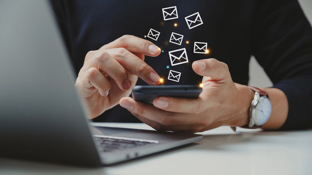 Person sitzt am Tisch und hält ein Handy in der Hand, aus welchem kleine Briefumschläge herausfliegen, vor ihr ist ein Laptop geöffnet